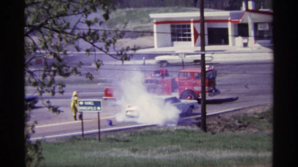 A firefighter in yellow gear stands near a smoking white sedan as a red fire truck waits along a Chicago roadside.
