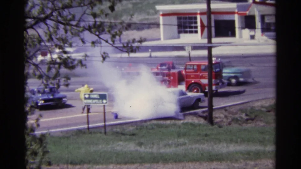 The smoking sedan is partly hidden by a dense white cloud while the fire truck and passing cars remain visible nearby.
