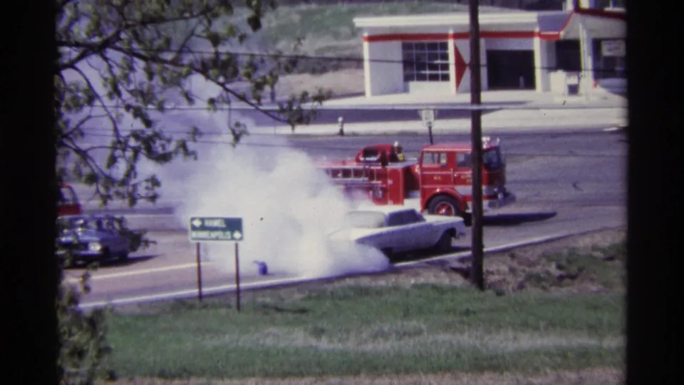 Final frame shows the white sedan still smoking on the roadside with a red fire truck, sign, and traffic in the background.