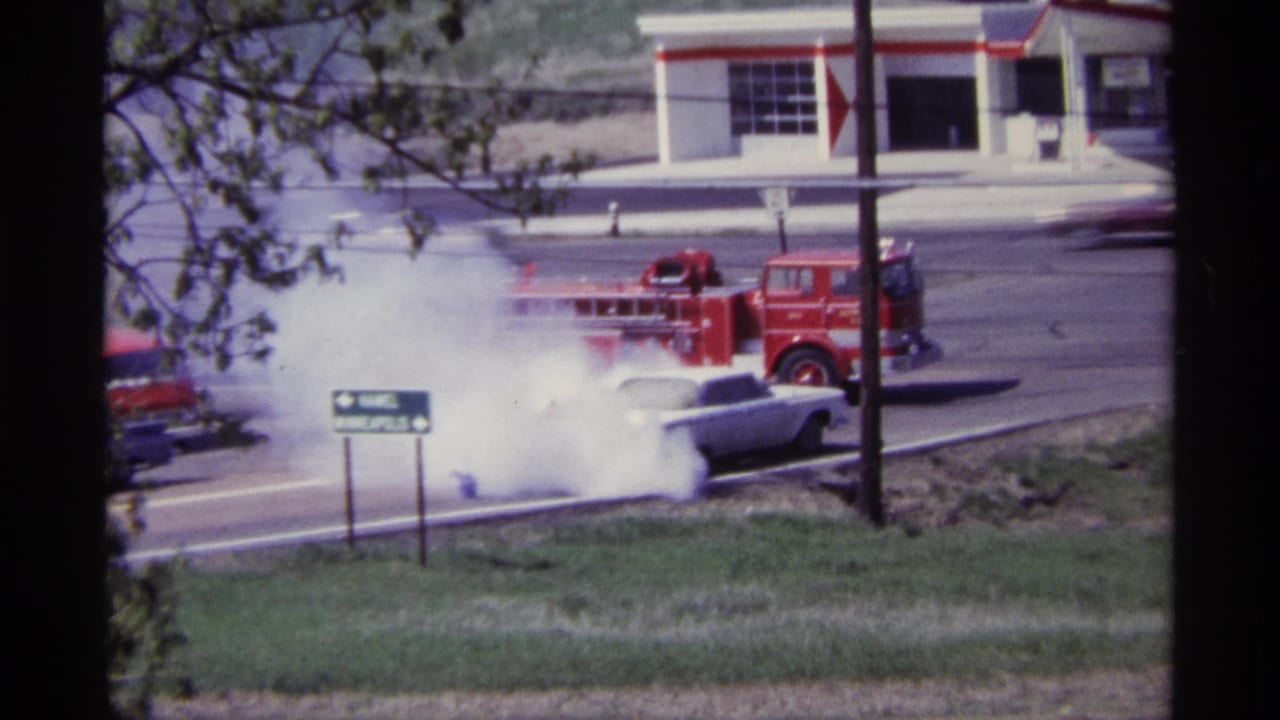 1968 Chicago home-movie frame showing a white car emitting heavy smoke near a red fire truck and roadside signage.
