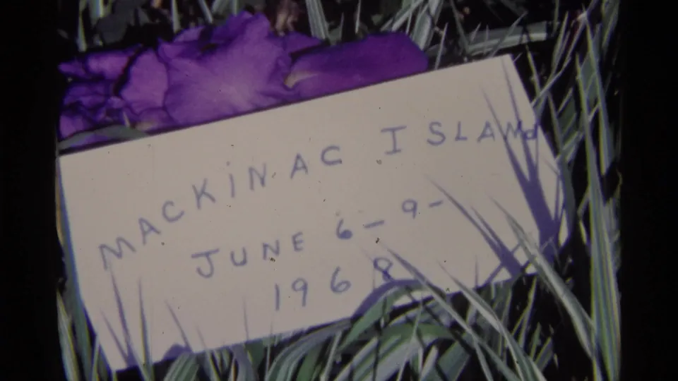 Home-movie close-up of a Mackinac Island June 1968 note framed by purple flowers and green grass.