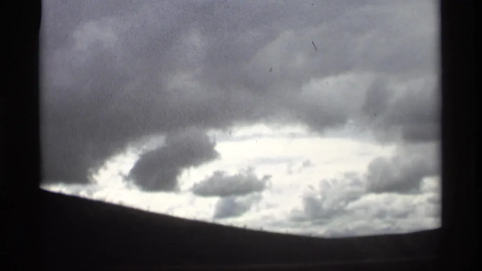1969 8mm frame showing a dark cloud mass over a bright break in the sky, viewed from moving travel footage.