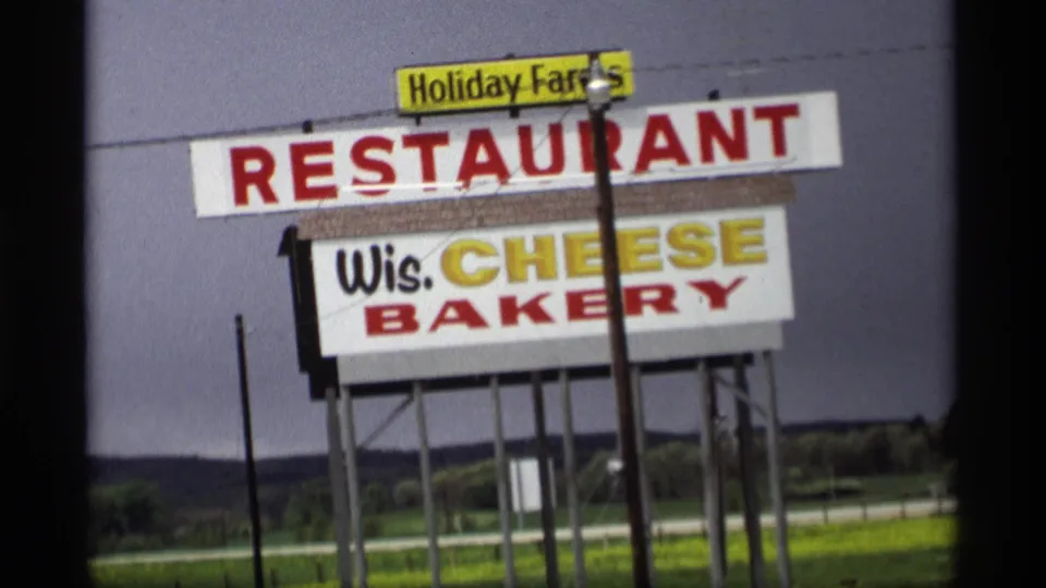 Closer 1969 Holland, Michigan view of the roadside Holiday Farms Restaurant and Wis. Cheese Bakery sign under a gray sky.
