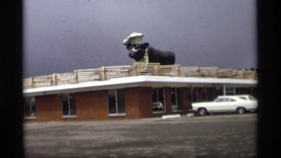 1969 Holland, Michigan frame showing a low roadside building with a giant black steer statue in a chef hat on the roof and a white 1960s car.