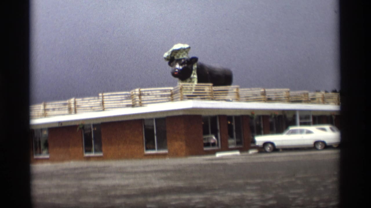 Brick motel exterior with a large animal chef statue on the roof and a white station wagon parked in front.