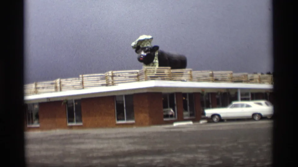 1969 Holland, Michigan home-movie view of a brick motel with a roof-mounted animal chef statue and a white station wagon outside.