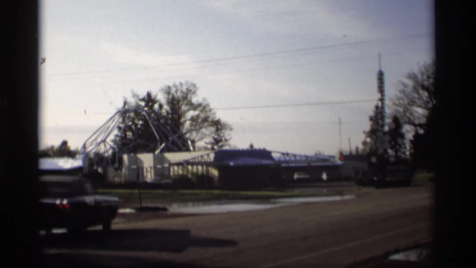 A dark car passes through the 1969 Holland, Michigan roadside view, with wet pavement, low buildings, wires, and trees behind it.