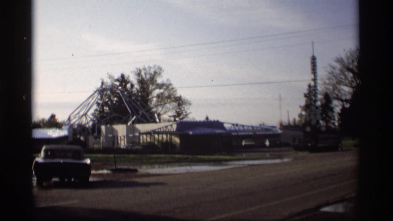 Roadside scene with a parked car, low building, metal framework, and distant tower.