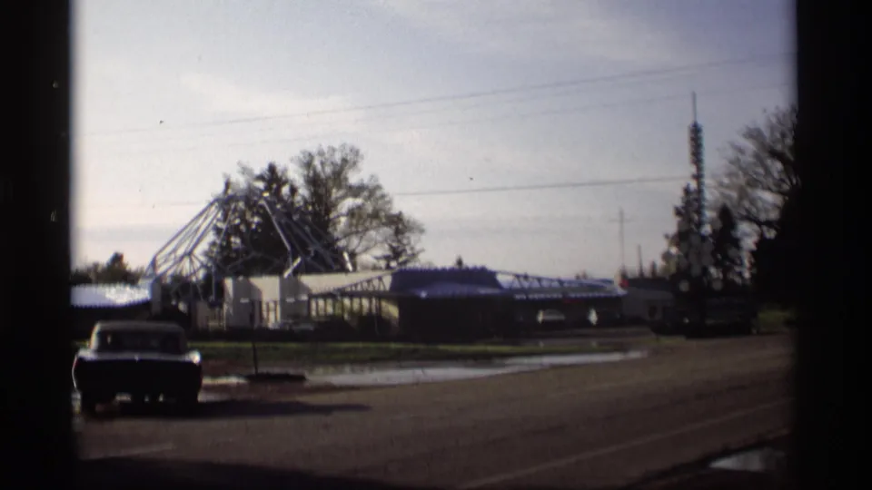 1969 Holland, Michigan roadside view with a parked car, low building, angular metal framework, and a tall tower in the distance.
