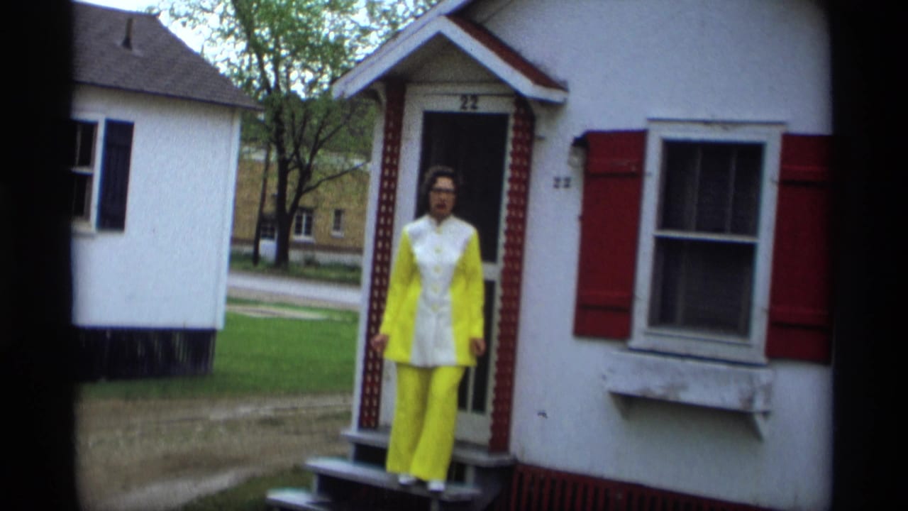 Woman in a yellow and white suit stands on the porch of a small white house with red shutters.