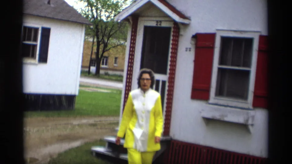 At the end of the clip, the woman walks toward the camera from the porch, with the white house and red shutters behind her.