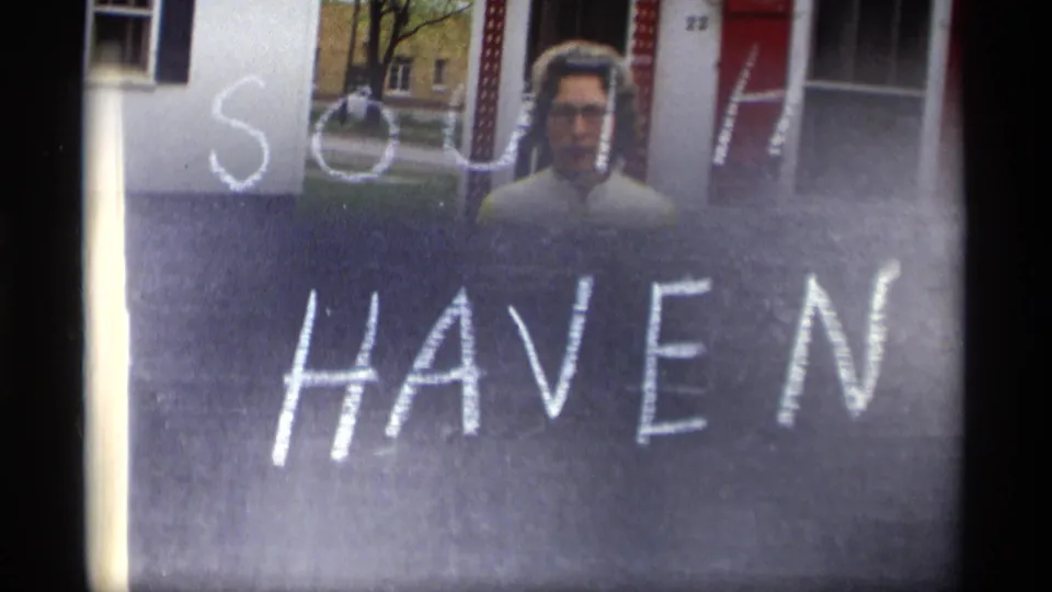 A woman appears behind a chalkboard marked SOUTH HAVEN, with part of a white house and red shutter visible behind her.