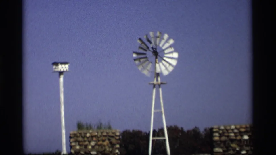 1969 Holland, Michigan 8mm frame with a white windmill at right, birdhouse pole at left, stonework, trees, and open sky.