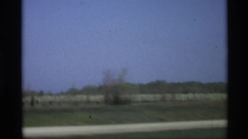 Moving 8mm footage shows a soft, blurred Holland, Michigan field with distant trees and a light roadside strip.