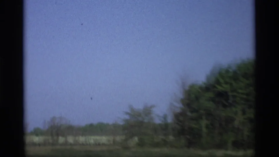 A 1969 8mm roadside view in Holland, Michigan with open sky, distant trees, and a darker tree mass passing at frame right.