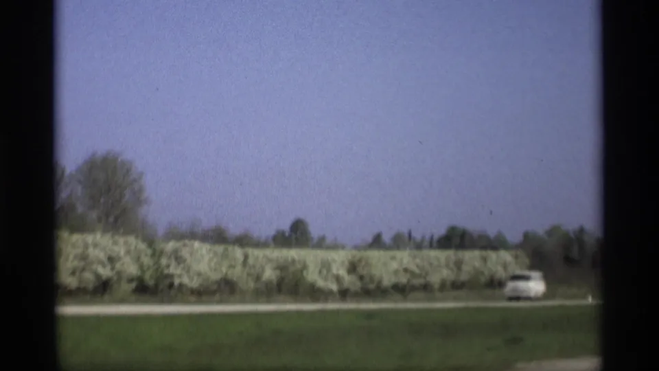 Home-movie view from 1969 near Holland, Michigan, showing a white car on a quiet road beside grass and flowering shrubs.