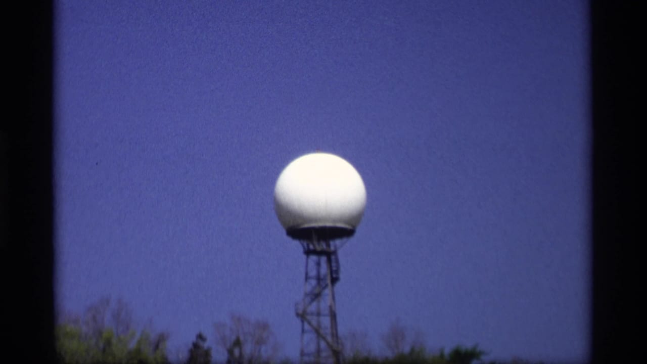 1969 Holland, Michigan 8mm frame showing a white radar dome atop a metal tower, with treetops below and deep blue sky behind it.