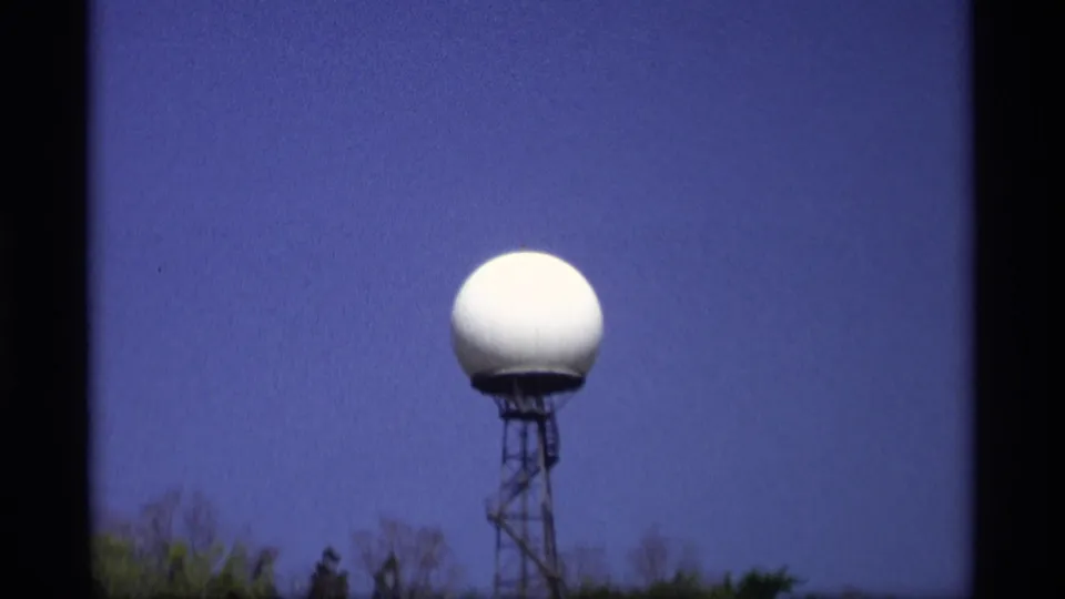 1969 Holland, Michigan 8mm frame showing a white radar dome atop a metal tower, with treetops below and deep blue sky behind it.