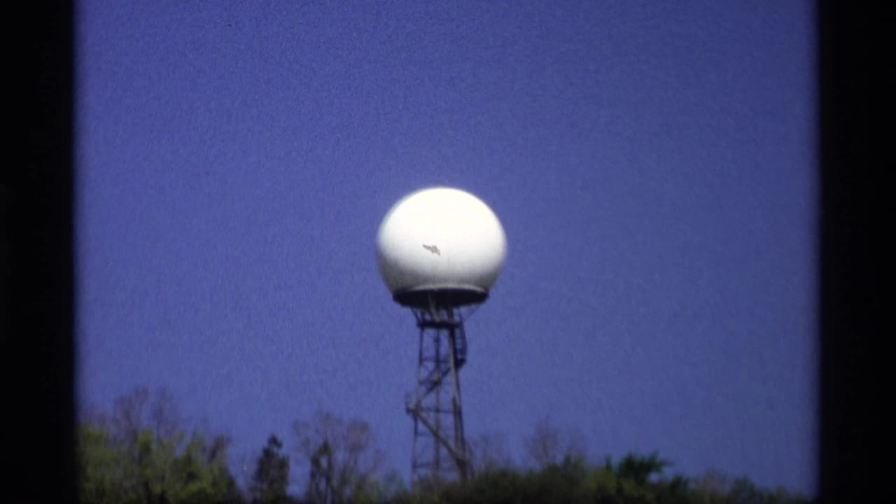 White radar dome on a tower with trees along the lower edge of the frame.