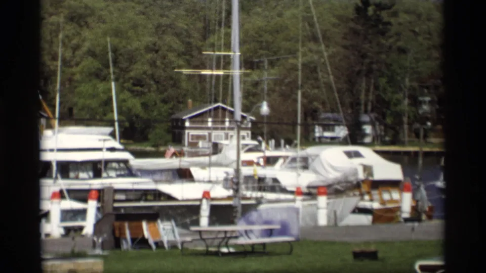 1969 Holland, Michigan marina view with motorboats, sailboat masts, a grassy foreground, wooded background, and a small dockside building.