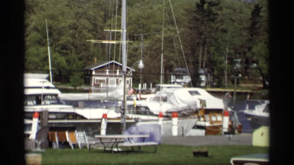 1969 Holland, Michigan 8mm marina frame showing covered boats, dock posts, tall masts, grass, and dense trees along the water.