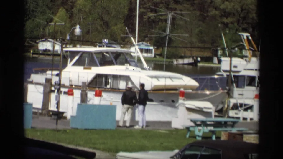 1969 marina scene in Holland, Michigan with a docked white yacht, two adults by the pier, a picnic table, grass, water, and trees.
