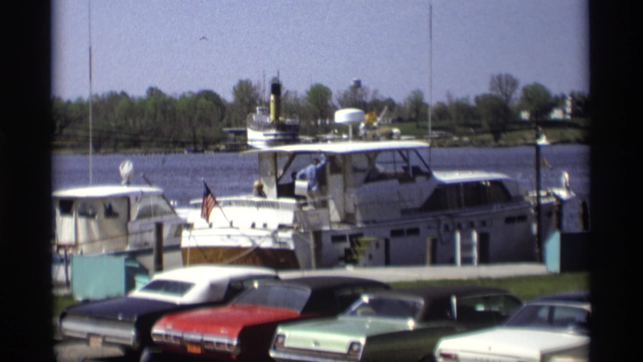 Classic cars parked beside a marina with a white motorboat on the water.