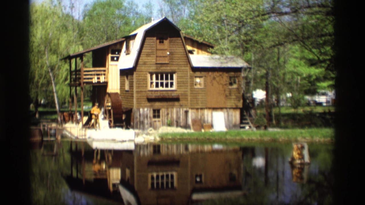 1969 Holland, Michigan 8mm frame showing a brown waterside mill structure, trees, calm water, reflection, and a small person near the wheel area.