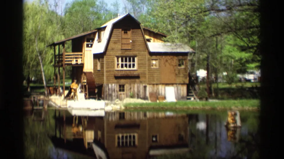 1969 Holland, Michigan 8mm frame showing a brown waterside mill structure, trees, calm water, reflection, and a small person near the wheel area.