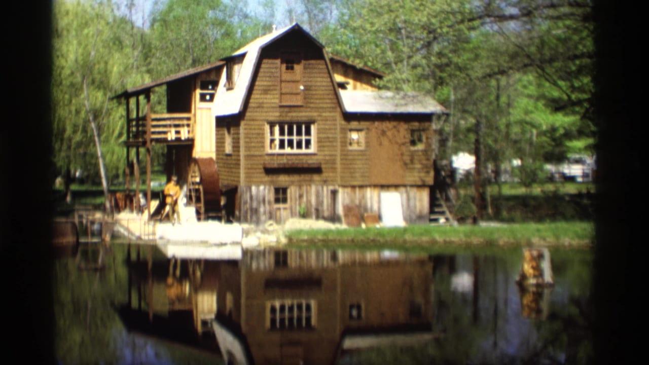 Wooden mill building and its reflection across calm water, framed by trees.