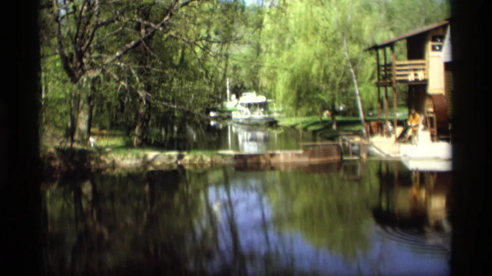 1969 Holland, Michigan frame looking along calm water toward a small boat, with trees, reflections, and the edge of the mill building at right.