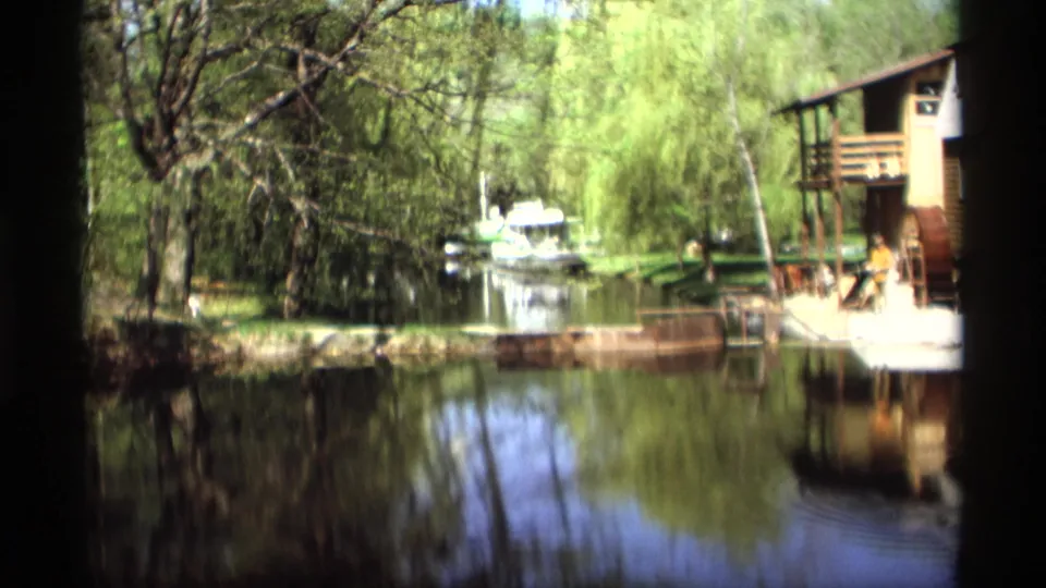 1969 Holland, Michigan 8mm frame showing reflective water, leafy trees, a small boat in the distance, and the right edge of the wooden mill structure.