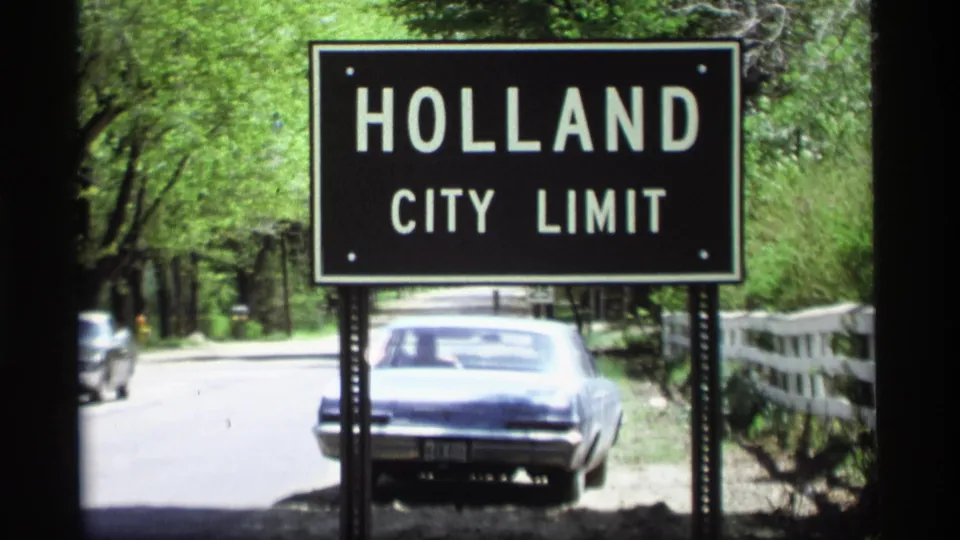 A black Holland city limit sign stands by a tree-lined road in Michigan, with a blue car, white fence, and 8mm film grain visible.