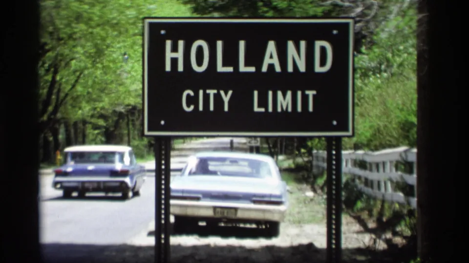 1969 Holland, Michigan frame with the city-limit sign foregrounding classic cars on a leafy roadside.