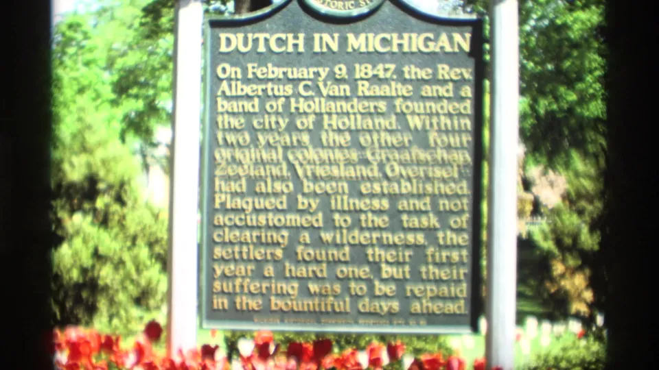 A public Dutch in Michigan historical marker is filmed in Holland, Michigan, with bright flowers and green trees around the sign.