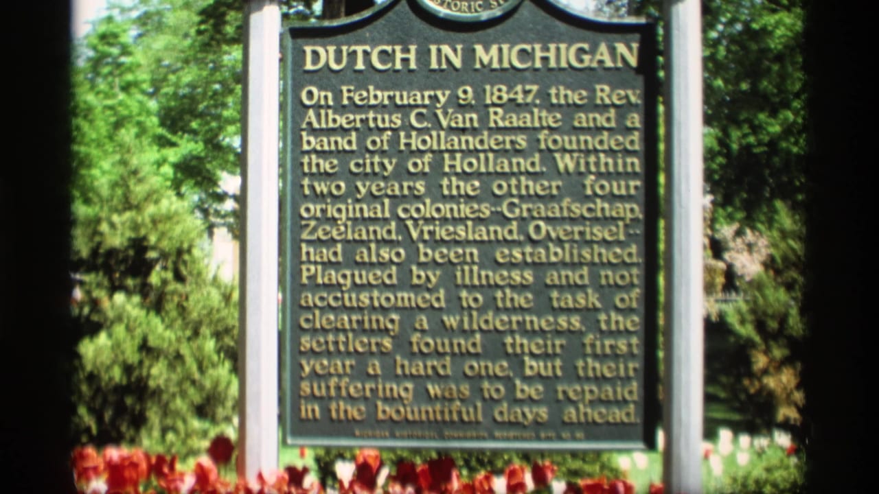 1969 Holland, Michigan 8mm frame showing the Dutch in Michigan marker with gold lettering, tulips, and trees in bright sun.