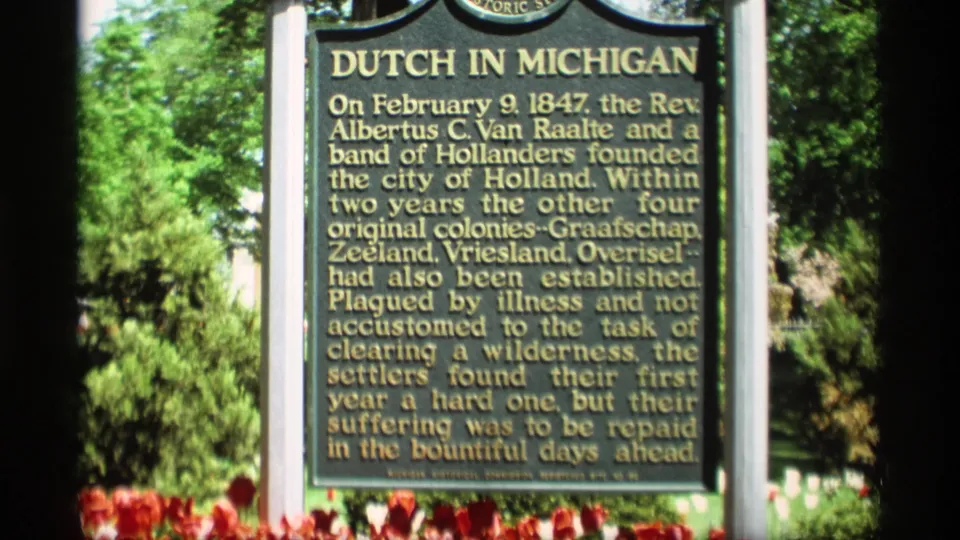 1969 Holland, Michigan 8mm frame showing the Dutch in Michigan marker with gold lettering, tulips, and trees in bright sun.