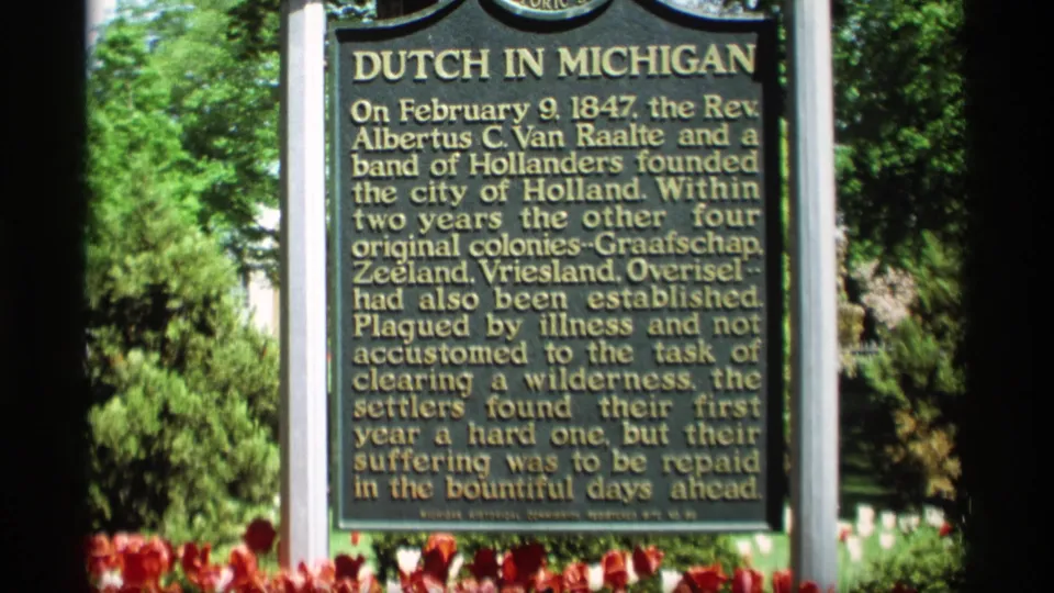 Close 8mm view of the Dutch in Michigan historical marker, with soft focus, gold lettering, tulips at the base, and trees nearby.