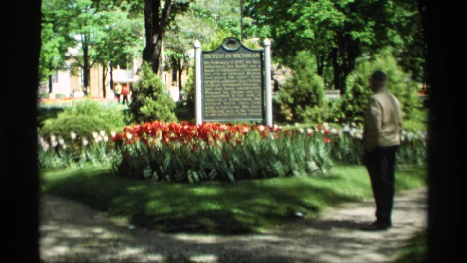 Wide 1969 Holland, Michigan frame with a man reading the Dutch in Michigan marker beside tulips, lawn, trees, and a paved path.