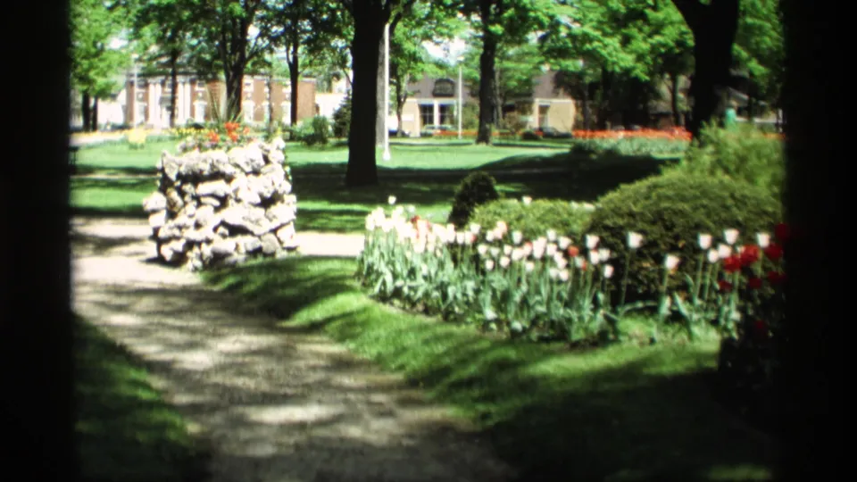 Holland, Michigan park view from 1969 showing a path, low stone structure, tulips, trees, lawn, and distant buildings.