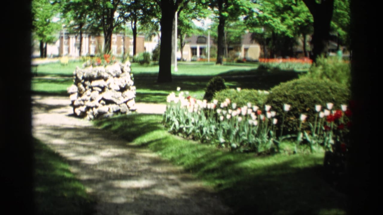 Shaded garden path with a stone pillar, tulip beds, trees, lawn, and a large house beyond.