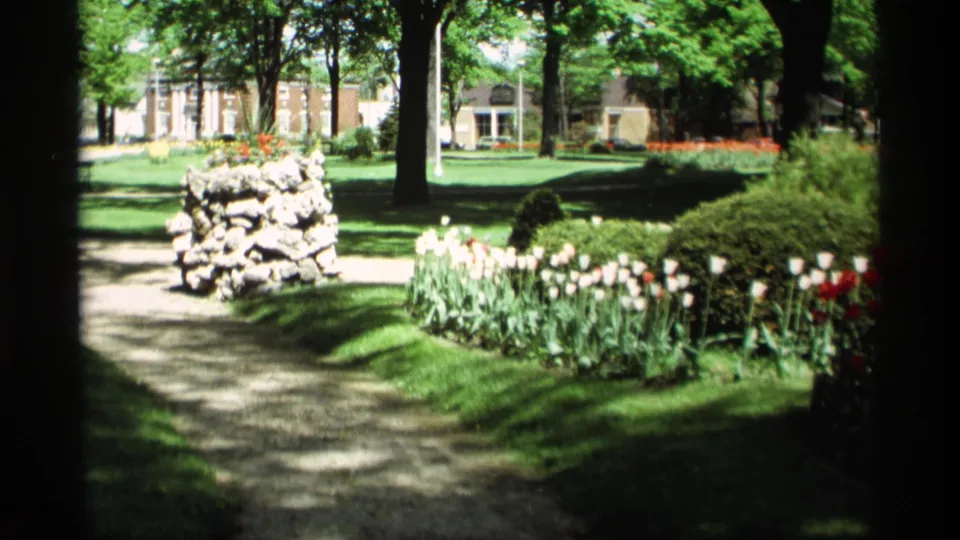 1969 Holland, Michigan garden scene with a gravel path, stone pillar, tulip beds, leafy trees, and a large house behind the lawn.