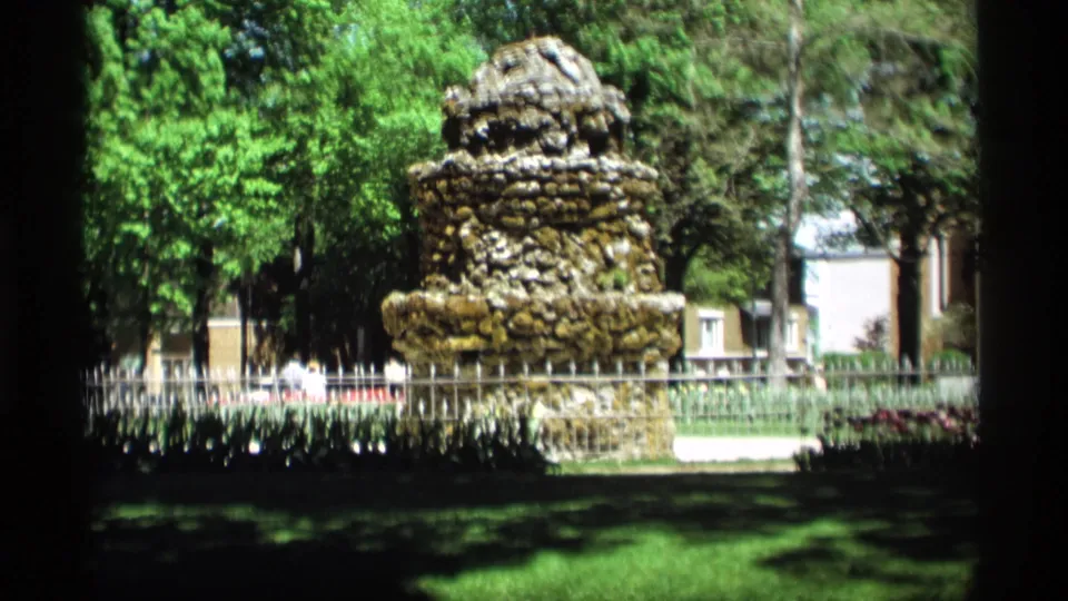 A Holland, Michigan park view shows a stone monument beyond a lawn, with trees, fencing, distant buildings, and possible distant visitors.
