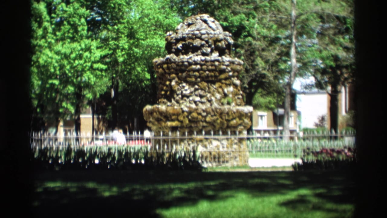 1969 home-movie view of a stone fountain in a Holland, Michigan park, framed by trees, lawn, and a low fence.