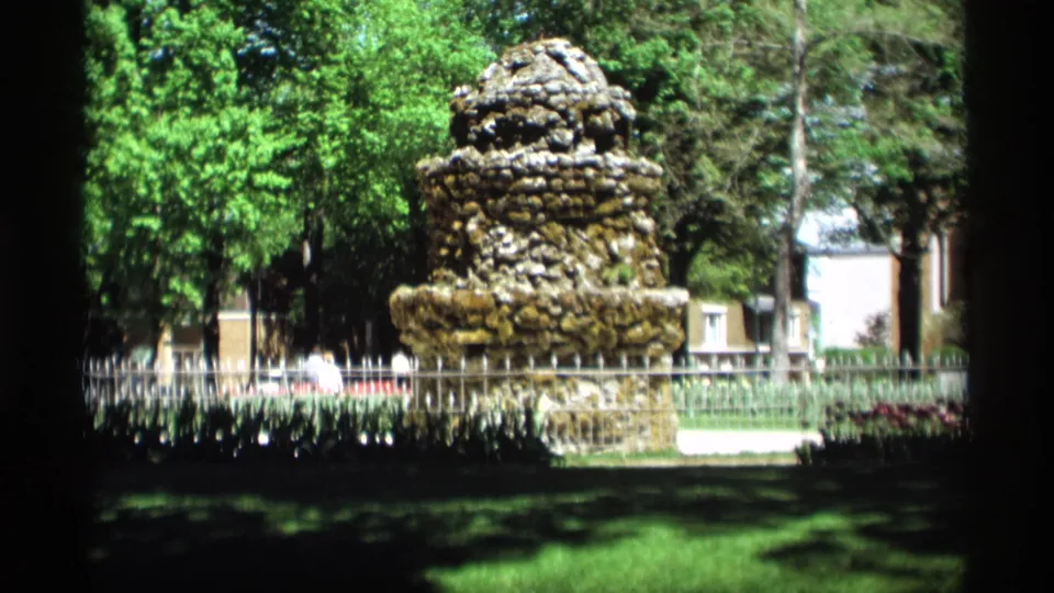 1969 home-movie view of a stone fountain in a Holland, Michigan park, framed by trees, lawn, and a low fence.