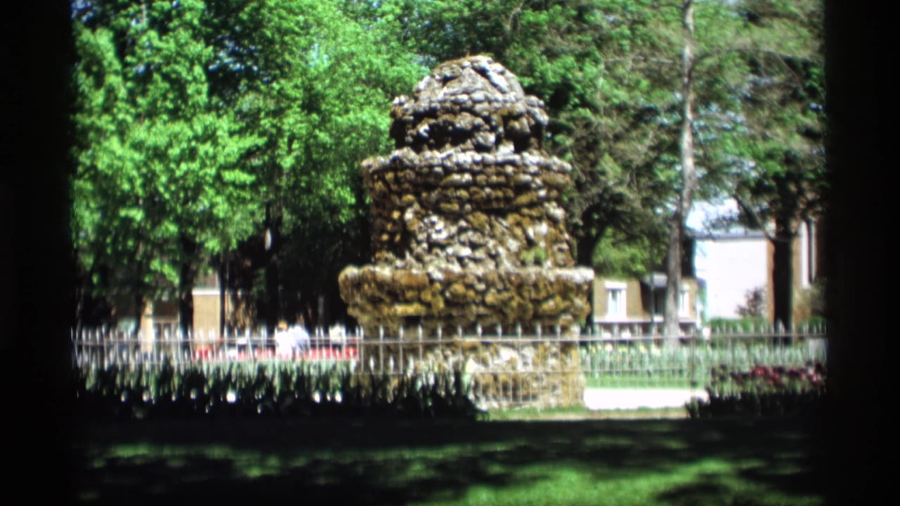 The Holland, Michigan park fountain remains centered, with distant pedestrians, trees, and buildings visible beyond the fence.