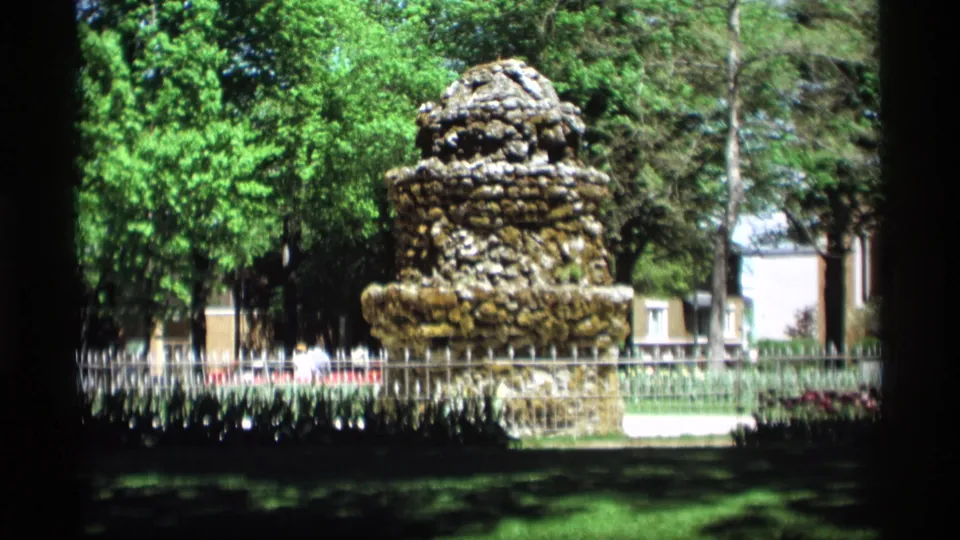 The Holland, Michigan park fountain remains centered, with distant pedestrians, trees, and buildings visible beyond the fence.