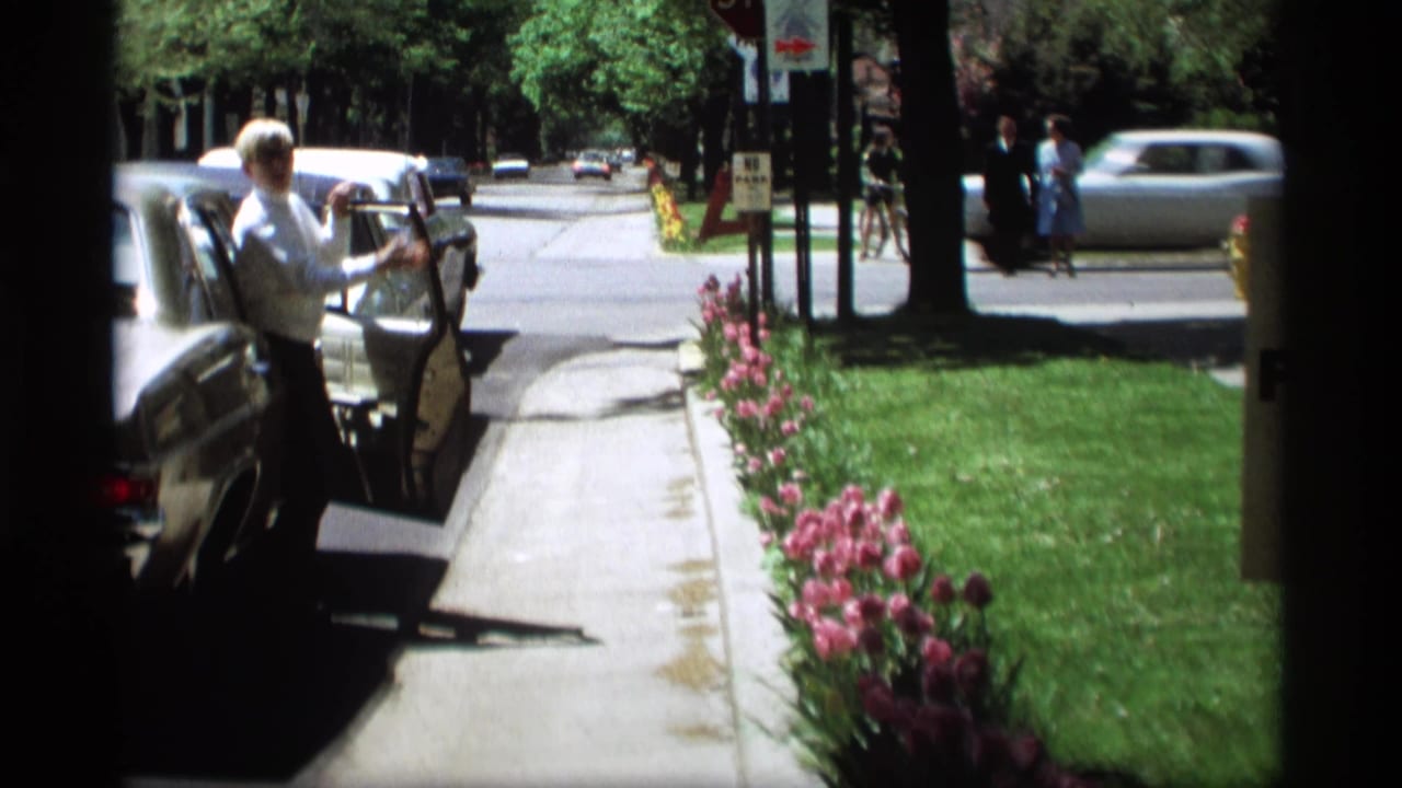 In Holland, Michigan home-movie footage from 1969, a man stands by an open car door beside tulips and a tree-lined street.