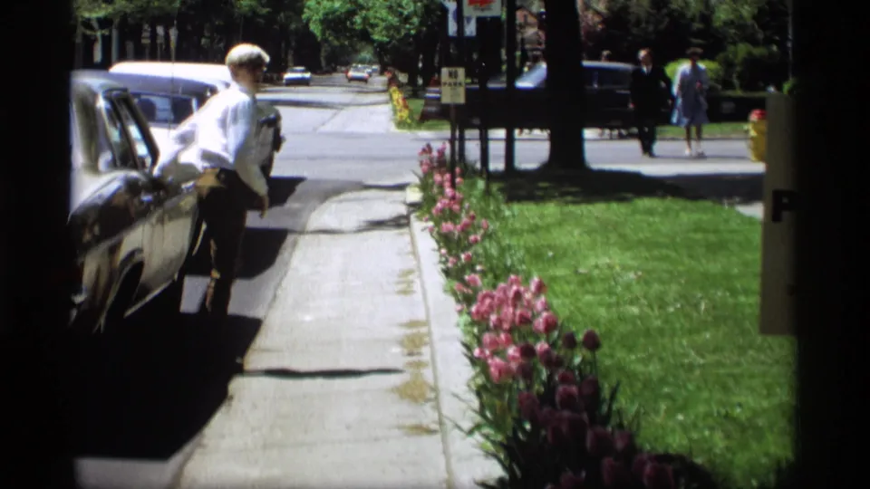 A man in a white shirt moves from the parked sedan toward the sidewalk in the 1969 Holland, Michigan street scene.