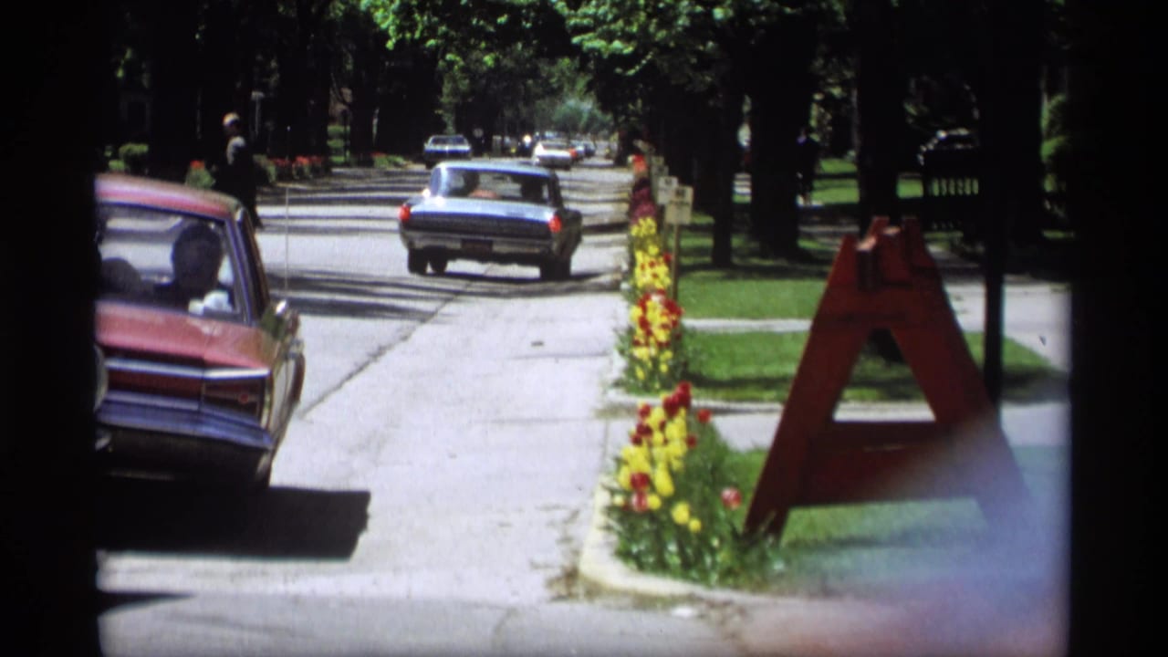 1969 Holland, Michigan 8mm frame showing cars on a shaded suburban street beside bright flower beds and a red A-frame sign.